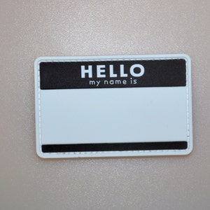 May include: A rectangular name tag with a white background and a black top section that reads "HELLO my name is" in white letters. The tag has a black border and is designed for personalization.