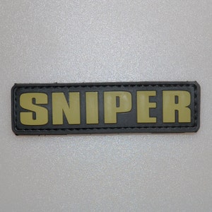 May include: A rectangular patch with the word "SNIPER" in olive green, raised letters. The letters are set against a black background with a stitched border. The patch is likely for military or tactical gear.