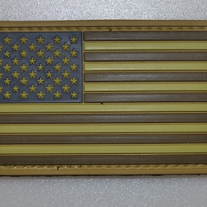 Może przedstawiać: Prostokątna naszywka z motywem flagi amerykańskiej. Flaga ma szare pole gwiazd, naprzemienne brązowe i beżowe paski oraz beżową obwódkę. Gwiazdy są żółte.