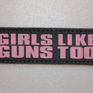 Könnte beinhalten: Schwarzer rechteckiger Aufnäher mit dem rosa Text "GIRLS LIKE GUNS TOO". Der Aufnäher hat einen genähten schwarzen Rand. Der Hintergrund ist eine helle, strukturierte Oberfläche.