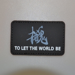 Könnte beinhalten: Ein rechteckiger schwarzer Aufnäher mit dem weißen Text "TO LET THE WORLD BE". Über dem Text befindet sich ein stilisierter, hellblauer japanischer Schriftzeichen. Der Aufnäher hat einen schwarzen Rand.
