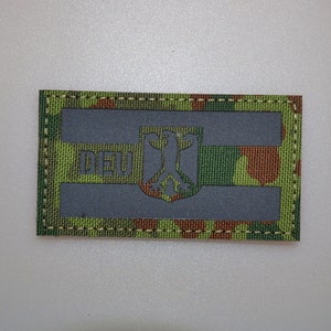 May include: A rectangular patch with a camouflage pattern in green, brown, and black. The patch features two horizontal gray bars and a gray emblem with a bird design. The letters "DEU" are visible in the upper left corner.