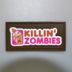 Puede incluir: Un parche rectangular marrón con las palabras "KILLIN' ZOMBIES" en naranja y rosa, con un gráfico de cabeza de zombi. El parche tiene un borde cosido.