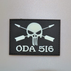 May include: Black rectangular patch featuring a white skull with crossed arrows and the text "ODA 516". The skull has a menacing expression. The patch has a black border and is likely made of a durable material.