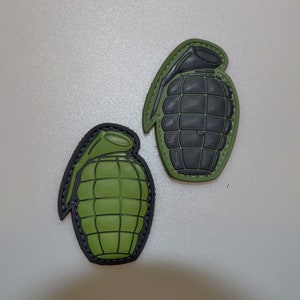 Puede incluir: Dos parches con forma de granada de mano. Uno es verde oliva con detalles negros, y el otro es negro con detalles verde oliva. Ambos tienen una apariencia texturizada y están sobre un fondo gris claro.