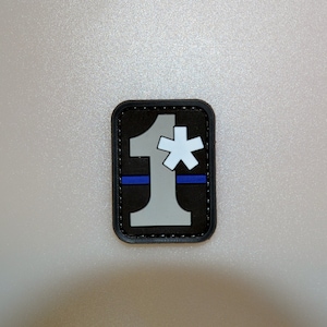May include: A rectangular patch with a black border. The patch features a gray number 1, a white asterisk, and a blue horizontal line on a black background. The patch is likely for tactical or uniform use.