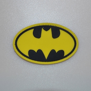 May include: A yellow and black Batman logo patch. The patch features the iconic bat symbol in black, set against a yellow oval background. The patch has a black border.