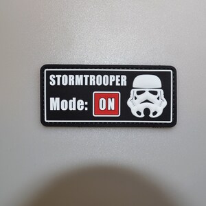 以下が含まれることがあります： 「STORMTROOPER Mode: ON」と白文字で書かれた黒い長方形のパッチ。右側には白いストームトルーパーのヘルメットのグラフィックがあります。「ON」という文字は赤いボックスに入っています。パッチは灰色の表面にあります。