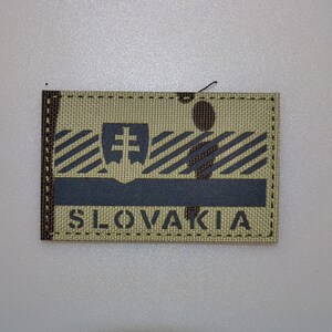 Pode incluir: Remendo retangular com a palavra "SLOVAKIA". O remendo tem um fundo bege com listras diagonais e uma faixa horizontal cinza escuro. Um emblema cinza escuro está acima da faixa horizontal.