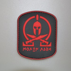 May include: A black and red patch featuring a Spartan helmet, crossed swords, and the Greek phrase "Molon Labe." The patch has a rectangular shape with rounded corners and a black border.