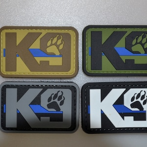 以下が含まれることがあります： 「K9」の文字と足跡がプリントされた長方形のパッチが4つ。パッチの色は、タン、オリーブグリーン、グレー、ブラック。各パッチには青いストライプと足跡のデザインが施されています。