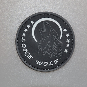 May include: A round, black patch featuring a howling wolf silhouette against a white crescent moon. The words "LONE WOLF" are arched around the design, with white stars bordering the top. The patch has a stitched border.
