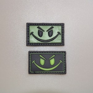 May include: Two rectangular patches with a smiling face design. The top patch has a green background with a black outline and black facial features. The bottom patch has a black background with a green outline and green facial features.