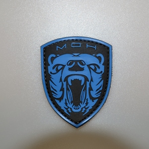 Op de afbeelding: Een zwart en blauw schildvormig embleem. Het embleem toont een brullende beer in blauw, met de letters "MOH" boven de beer. Het embleem heeft een zwarte rand en staat op een lichtgrijze achtergrond.