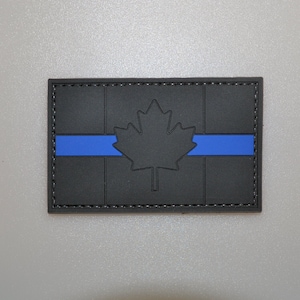 May include: A rectangular patch with a black background and a blue horizontal stripe. A black maple leaf is centered on the patch. The patch has a black border and is likely made of rubber or a similar material.