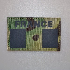 Puede incluir: Un parche rectangular con la palabra "FRANCE" en letras negras. El fondo presenta un patrón de camuflaje en tonos verdes, marrones y beige. Dos cuadrados negros están debajo de la palabra.