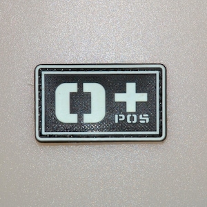 Könnte beinhalten: Ein rechteckiger Aufnäher mit schwarzem Rand und strukturiertem Hintergrund. Der Aufnäher zeigt den Text "[]+ POS" in Weiß, mit dem Symbol "[]" und einem Pluszeichen. Das Design ist schlicht und markant.