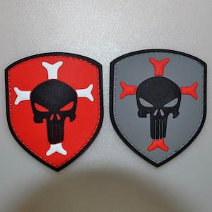 Puede incluir: Dos parches en forma de escudo con un diseño de calavera. Uno tiene un fondo rojo con una calavera negra y huesos cruzados blancos. El otro tiene un fondo gris con una calavera negra y huesos cruzados rojos. Ambos tienen un borde negro.