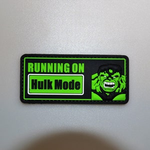 Puede incluir: Un parche rectangular con el texto "RUNNING ON Hulk Mode" en blanco y verde. El parche presenta una ilustración de Hulk, un superhéroe de piel verde, con una expresión enfadada. El parche tiene un borde negro.