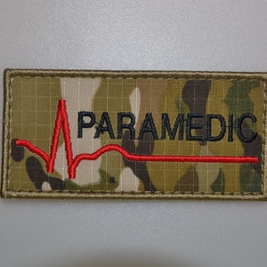 Könnte beinhalten: Gestickter Aufnäher mit dem Wort "PARAMEDIC" in Schwarz, über einer roten Herzschlaglinie. Der Aufnäher hat einen Tarnhintergrund und einen beigen Rand. Für medizinisches Personal.