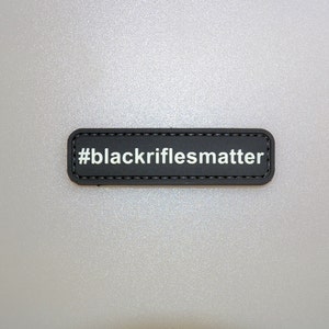 May include: A black rectangular patch with the text "#blackriflesmatter" in white. The patch has a stitched border and is set against a light gray background. The patch is a simple, yet bold statement.