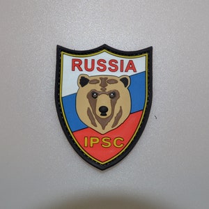 Op de afbeelding: Een schildvormige patch met een berenkop en het woord "RUSSIA" in het rood. De patch heeft een wit, blauw en rood kleurenschema, met de tekst "IPSC" in het rood aan de onderkant. De patch is zwart omlijnd.