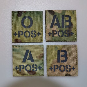 May include: Four square patches with a camouflage pattern. Each patch displays a different blood type: O, AB, A, and B, along with the text '+POS+' in black stencil font. The patches are likely for military or medical use.