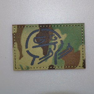 May include: Rectangular camouflage patch with a dark gray graphic design. The patch features a green, brown, and tan camouflage pattern. The graphic appears to be a stylized face or symbol. The edges are stitched with black thread.