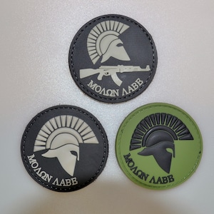 Può includere: Tre toppe rotonde con un disegno di elmo spartano. Una è nera con un elmo bianco e un fucile AK-47, e il testo "MOLON LABE". Le altre due sono nere e verdi con un elmo spartano e il testo "MOLON LABE".