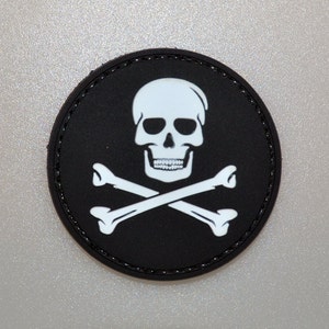 Könnte beinhalten: Ein runder, schwarzer Aufnäher mit einem weißen Totenkopf und gekreuzten Knochen. Der Totenkopf hat detaillierte Augenhöhlen und Zähne. Der Aufnäher hat einen genähten Rand, und das Design ist zentriert.