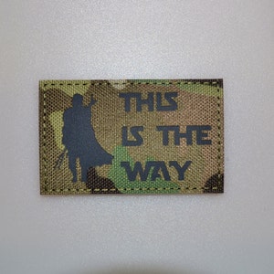 May include: A rectangular camouflage patch with a black silhouette of a figure and the text "THIS IS THE WAY". The patch has a stitched border. The text is in block letters.