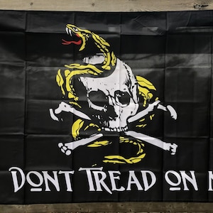 Pode incluir: Bandeira preta com uma caveira branca, ossos cruzados e uma cobra amarela enrolada em volta da caveira. As palavras "DONT TREAD ON ME" estão impressas em branco abaixo.