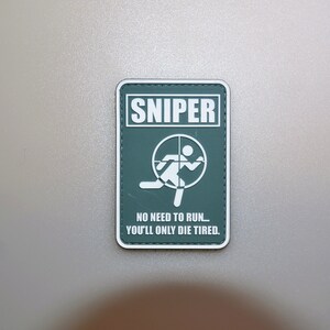 Op de afbeelding: Een rechthoekige patch met het woord "SNIPER" in witte blokletters boven een afbeelding van een persoon in een vizier. Onder de afbeelding staat de tekst: "NO NEED TO RUN... YOU'LL ONLY DIE TIRED." De patch heeft een groene achtergrond en een witte rand.