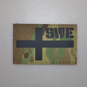Puede incluir: Un parche rectangular con un patrón de camuflaje. Una cruz negra está centrada, con las letras "SWE" encima. El parche es probablemente para uso militar o táctico.