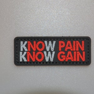Op de afbeelding: Een rechthoekige zwarte patch met de tekst "KNOW PAIN KNOW GAIN" in witte en rode letters. De patch heeft een zwarte rand en is waarschijnlijk gemaakt van een duurzaam materiaal.