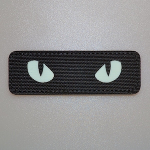 Op de afbeelding: Een rechthoekige zwarte patch met een getextureerd oppervlak. Twee lichtgroene, gestileerde oogvormen zijn gecentreerd op de patch. De patch is waarschijnlijk een opstrijk- of opnaai-applicatie.