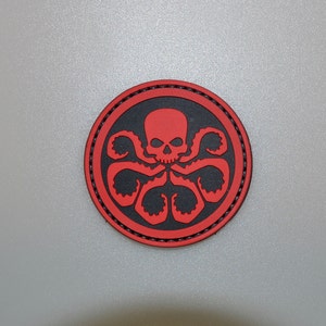 Puede incluir: Un parche redondo con un diseño de calavera roja y tentáculos de pulpo sobre un fondo negro. El diseño está enmarcado por un borde rojo con costuras negras. Adecuado para ropa o accesorios.