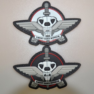 May include: Two tactical patches with a skull, knife, wings, and the Latin text "Timent Canis Morsum." The patches have a black border, one with a white background and the other with a black background, both with grey and red detailing.