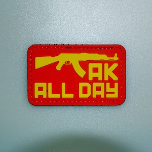 Può includere: Una toppa rettangolare rossa con una silhouette gialla di un fucile AK-47 e la scritta "AK ALL DAY" in lettere gialle. La toppa ha un bordo nero ed è probabilmente fatta di tessuto.