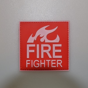 May include: A red square patch with a pink border and a white design. The design features a flame and the words "FIRE FIGHTER" in bold white letters. The patch is likely for a firefighter's uniform or gear.