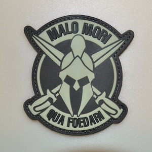 Peut inclure: Un écusson noir et vert avec un casque spartiate, des épées croisées et la phrase latine "MALO MORI QUAM FOEDARI". L'écusson a un design circulaire avec une bordure cousue.