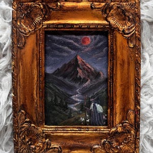 May include: A framed painting depicting a mountain landscape under a starry night sky with a red moon. The painting is in a gold ornate frame. The artwork features a figure with a staff in the foreground, and a river winding through the valley.