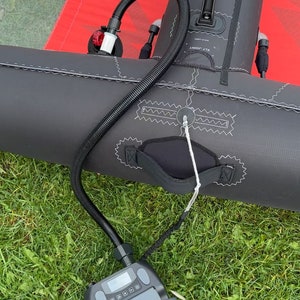 May include: A black and red inflatable wing with a pump and hose on green grass. The wing has a handle and is connected to a gray pump with a digital display. A sneaker is visible in the lower left corner.