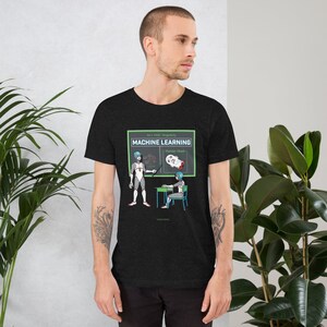 May include: Dark gray t-shirt featuring a graphic of a machine learning lesson. The design includes a robot teacher, a student, and a chalkboard with the text "MACHINE LEARNING" and "Human Head". The shirt has a crew neck and short sleeves.