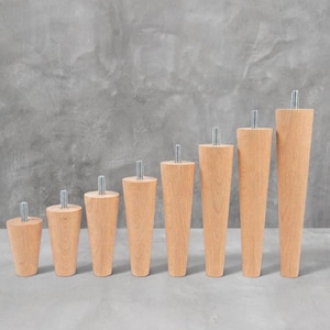 Könnte beinhalten: Ein Set aus acht hellen Holzmöbelbeinen, die in aufsteigender Höhe angeordnet sind. Jedes konische Bein hat eine glatte Oberfläche mit natürlicher Holzmaserung und einen silberfarbenen Metallgewindebolzen an der flachen Oberseite.
