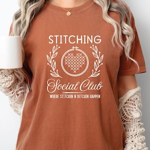 May include: A rust-colored t-shirt with a white graphic that says "Stitching Social Club" with a stylized heart and wreath design. The text below the graphic says "Where Stitchin' & Bitchen' Happen."