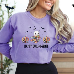 May include: A lilac-colored sweatshirt with a Halloween-themed graphic. The design features a ghost character wearing a plaid scarf, flanked by two patchwork pumpkins, and the text "HAPPY QUILT-O-WEEN" below. Black bat silhouettes are scattered above.