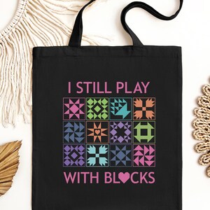 May include: Black tote bag with the text "I still play with blocks" in pink and colorful quilt block designs.