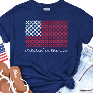 May include: Navy blue t-shirt with a stitched American flag design. The flag features red and blue cross-stitch patterns, with the text "stitchin' in the usa" in white script. The shirt is paired with denim shorts and sandals.