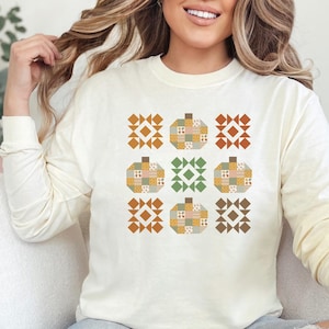 May include: Cream-colored long-sleeve shirt featuring a patchwork pumpkin design. The pumpkins are in various colors, including orange, green, and yellow, with geometric patterns. The shirt is a casual style.